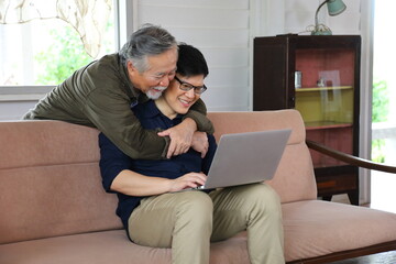 Senior Asian man in retirement age hugging his son from behind while he is working from home using laptop with copy space