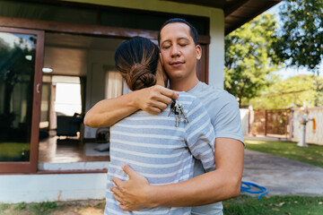 Portrait of 30s happy young adult Asian couple embracing while holding a house key. Husband and wife real estate owners hugging in front of their homes. Housing investment achievement concept