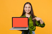 Smiling Young Brunette Asian Woman In Basic Green Shirt Standing Pointing Index Finger On Laptop Pc Computer With Blank Empty Screen Mock Up Copy Space Isolated On Yellow Background, Studio Portrait.