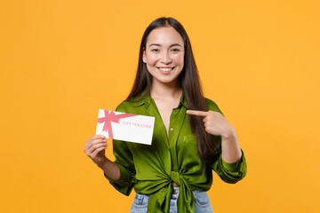 Wall Mural - Smiling beautiful young brunette asian woman wearing basic green shirt standing pointing index finger on gift certificate looking camera isolated on bright yellow colour background, studio portrait.