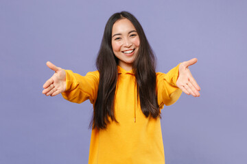 Wall Mural - Smiling joyful beautiful attractive young brunette asian woman 20s in casual yellow hoodie standing reach out stretch hands looking camera isolated on pastel violet colour background studio portrait.