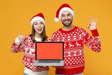 Funny young Santa couple friends man woman in Christmas hat hold laptop computer with blank empty screen showing OK gesture thumb up isolated on yellow background. Happy New Year celebration concept.