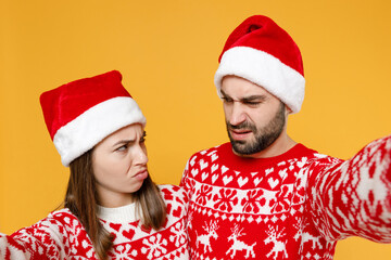 Wall Mural - Close up offended young Santa couple friends man woman in red sweater, Christmas hat doing selfie shot on mobile phone isolated on yellow background. Happy New Year celebration merry holiday concept.