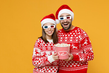 Wall Mural - Excited young Santa couple friends man woman in sweater, Christmas hat 3d glasses watching movie film holding bucket of popcorn isolated on yellow background. Happy New Year celebration merry concept.