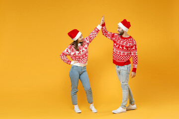 Wall Mural - Full length of cheerful young Santa couple friends man woman in sweater, Christmas hat giving high five looking at each other isolated on yellow background. Happy New Year celebration holiday concept.