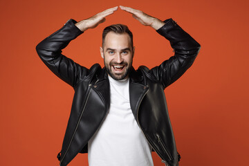 Excited young bearded man 20s wearing basic white t-shirt black leather jacket standing holding hands above head like roof of house looking camera isolated on orange colour background studio portrait.