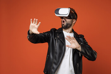 Poster - Shocked young bearded man 20s wearing white t-shirt black leather jacket standing watching in vr headset gadget touch something like push click on button isolated on orange background studio portrait.