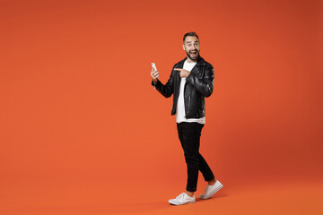 Wall Mural - Full length side view of shocked young bearded man in basic white t-shirt black leather jacket standing pointing index finger on mobile cell phone isolated on orange colour background studio portrait.