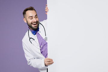 Wall Mural - Excited young bearded doctor man wearing white medical gown hold big white empty blank billboard isolated on violet colour background studio portrait. Healthcare personnel health medicine concept.