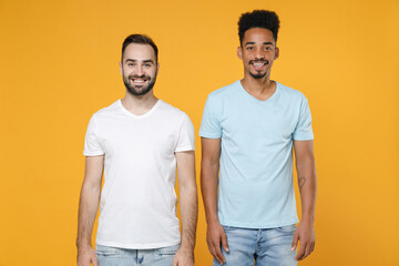 Sticker - Smiling cheerful young two friends european african american men 20s wearing white blue casual t-shirts standing and looking camera isolated on bright yellow colour wall background studio portrait.