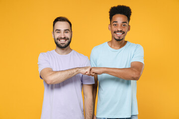 Cheerful smiling young friends european african american men 20s wearing casual violet blue t-shirts giving fists bump looking camera isolated on bright yellow colour wall background studio portrait.