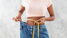 Black Woman In Old Big Jeans Measuring Her Waist, Showing Results Of Slimming Diet Or Liposuction, Grey Background
