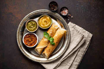 Wall Mural - Traditional Indian rice pancakes Dosa with different dips chutney, seasonings on rustic metal plate on dark brown stone background table. Quick meal or vegetarian snack of South India, top view