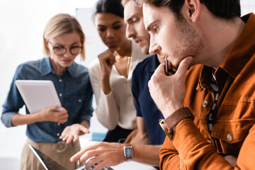 thoughtful multiethnic businesspeople pointing with hands at laptop while thinking together on blurred background