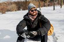 Winter Lifestyle Portrait Of Handsome Man With Beard And Backpack  Sitting On Snow In Winter Forest. Holidays And Travel Concept.