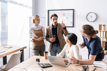 Wall Mural - Multicultural office workers looking at excited colleague with idea gesture while standing in office near workplaces