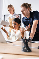 Wall Mural - Offended multicultural office workers gesturing while looking at laptop at workplace with blurred pens holder on foreground