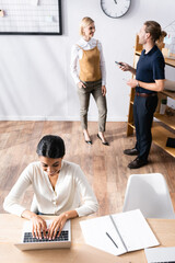 Wall Mural - Overhead view of happy african american woman typing on laptop while sitting at desk with office workers talking on background