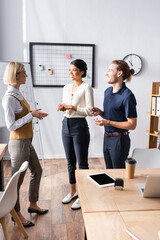 Wall Mural - Smiling multicultural office workers talking while standing near workplace