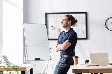 Wall Mural - Confident businessman with crossed arms looking away while leaning on table in office