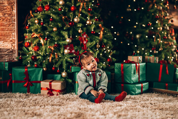 Wall Mural - Little girl sitting in front of a decorated Christmas tree. Against the background of a Christmas tree with boxes in green packaging with a bow. Christmas mood