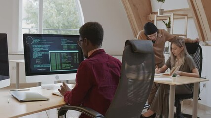 Back view of young African male coder wearing casual clothes sitting at workplace and developing program code on computer while his colleagues talking