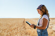 © Alvaro Sanchez/ADDICTIVE STOCK - Side view of positive focused female farmer in overalls standing in wheat field in countryside and browsing tablet