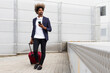 © David Munoz/ADDICTIVE STOCK - Black man in a suit with a wireless headphones around his neck and a suitcase walking while using the mobile outdoors