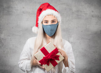 Young beautiful blond woman wears christmas santa claus hat - cap and holding red gift box - present with ribbon, isolated on gray background. A Corona - Covid 19 Christmas concept