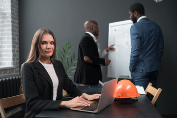 Wall Mural - Caucasian young confident businesswoman sitting at the office table with group of colleagues in the background, working on laptop computer