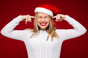 Wall Mural - Stock photo of pretty woman with blonde hair in white sweater and Santa hat plugging her ears with hands