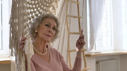 Calm pensioner woman wearing nice white pearl jewelry and pink jacket sits and swings in hammock by windows closeup slow motion