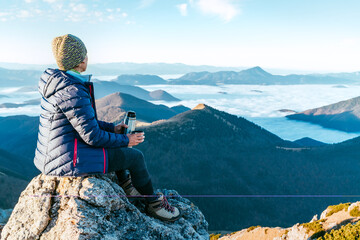 Wall Mural - Young hiker woman sitting on the mountain summit cliff and enjoying mountains valley covered with clouds view. Successful summit climbing concept image.