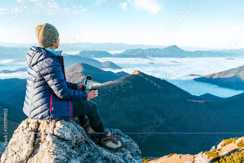 Fotomural Young hiker woman sitting on the mountain summit cliff and enjoying mountains valley covered with clouds view