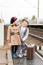 Joyful Children, A Boy And A Girl Dressed In Vintage Clothes .. Photo At The Station Near The Railway Tracks. A Retro Suitcase Stands Nearby