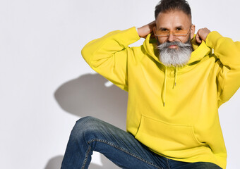 Brutal bearded smiling middle aged man in bright yellow hoodie and jeans sitting and putting on hood over white wall background. Stylish bright comfortable casul fashion for men concept