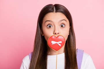 Sticker - Portrait of astonished kid girl cover mouth with heart shape candy wear purple white outfit isolated on pink color background