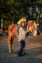 A Young Beautiful Brunette In A Plaid Shirt Holds The Reins Of A Brown Horse, On Which Sits A Cute Baby 4 Years Old