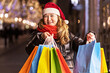 © Светлана Густова - A woman with long hair and a Santa hat near the window of a city store with purchases in colorful, paper bags. New year's shopping