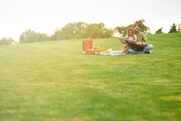 Full length shot of happy little girl sitting with her loving father on a green grass in park and learning how to play guitar