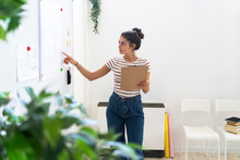 Young Businesswoman Pointing At Whiteboard While Holding Clipboard In Creative Workplace
