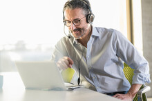 Mature businessman wearing headphones using laptop at desk in office
