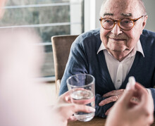 Woman Holding Pill And Glass Of Water For Smiling Senior Man