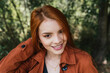 © Daniel Sierralta/Westend61 - Smiling beautiful young redhead woman wearing brown jacket at park