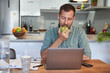 © victor espadas gonzalez/Westend61 - Handsome male freelancer eating fresh apple while working at home with laptop