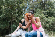 Cheerful Young Woman Taking Selfie With Redhead Female Friend Sitting At Park On Sunny Day