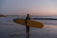 Surfer Holding Surf Board Standing On Shore At Sunset