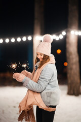 Wall Mural - Cheerful young woman celebrating holding sparkles in the winter forest. Festive garland lights. Christmas, new year.