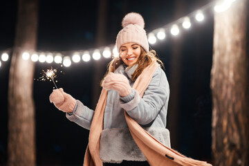 Wall Mural - Cheerful young woman celebrating holding sparkles in the winter forest. Festive garland lights. Christmas, new year.