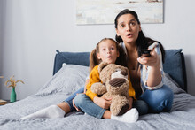 Excited Mother And Daughter Watching Tv While Sitting On Bed With Teddy Bear